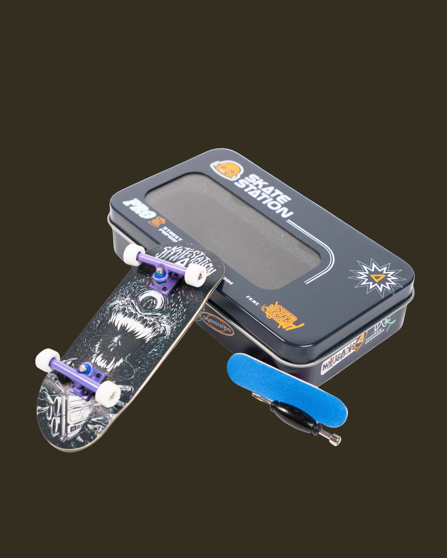 fingerboard with trucks, a metal carrying case, and a blue tool in a black tin labeled 'Skatestation' on a white background.